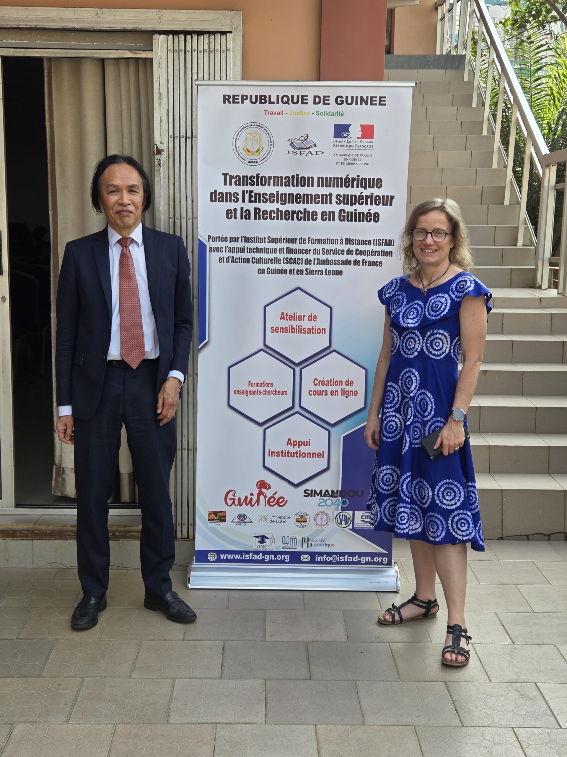 Deux représentants de L'Université Numérique posant devant une affiche ayant pour titre : transformation numérique dans l'Enseignement supérieur et le Recherche en Guinée.